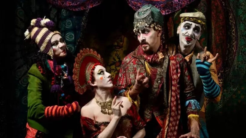 Grupo de actores caracterizados con vestuario teatral colorido y maquillaje escénico en una escena dramática.
