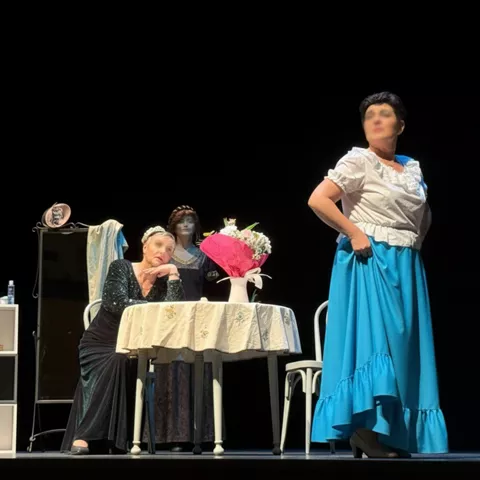 Escena con dos personas junto a una mesa, una sentada y otra de pie con falda azul y fondo negro.