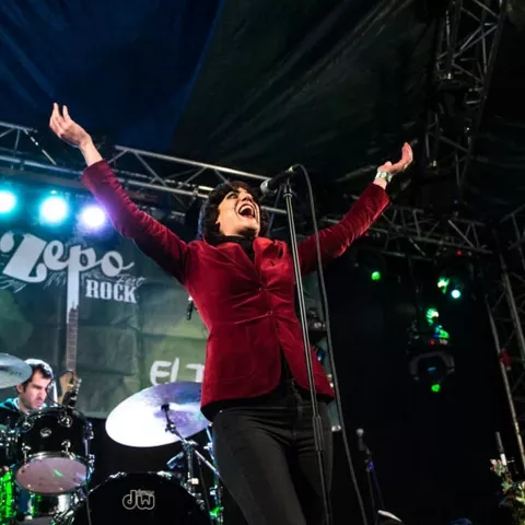 Cantante con los brazos en alto junto a su banda tocando en un escenario con luces de colores.