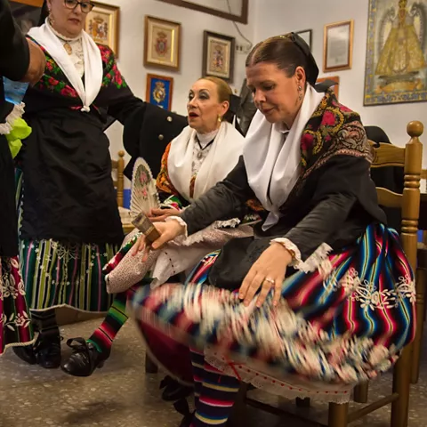 Preparativos de indumentaria tradicional en una sala con personas sentadas y de pie.