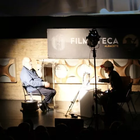 Charla en un auditorio con dos personas sentadas frente al público, iluminadas por focos y equipo de grabación.