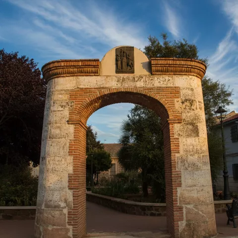Arco conmemorativo de piedra y ladrillo en una plaza ajardinada de Granátula de Calatrava (Ciudad Real).