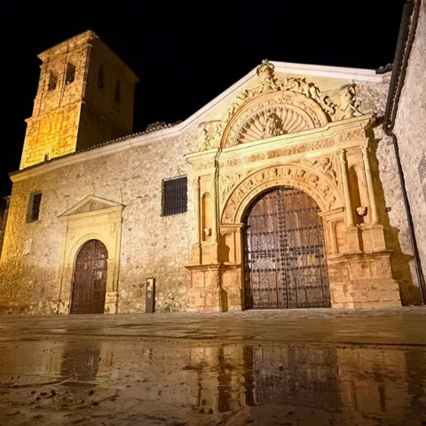 Fachada de iglesia de piedra iluminada de noche con torre campanario y suelo mojado en primer plano.