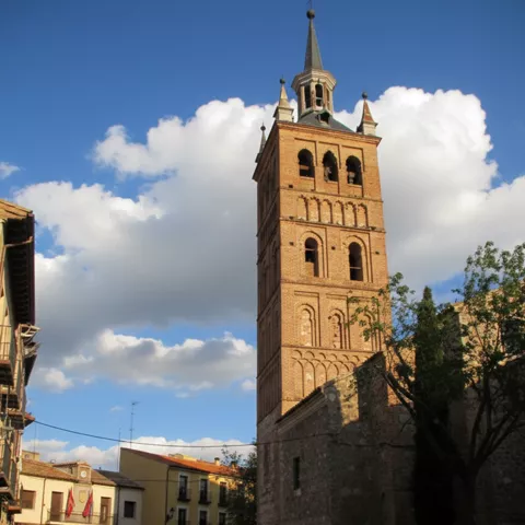 Torre campanario junto a plaza con edificios bajos y cielo parcialmente nublado.
