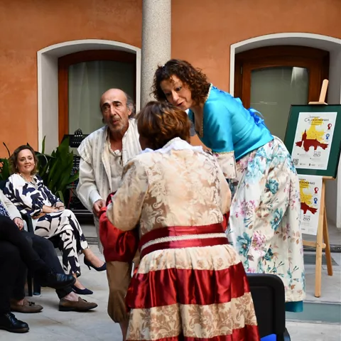 Representación teatral en un patio con actores vestidos de época ante público sentado.