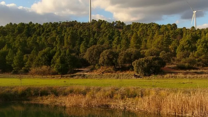 Laguna y paisaje forestal en Masegoso con aerogeneradores sobre la sierra bajo un cielo parcialmente nublado.