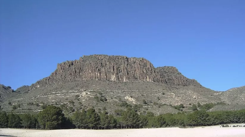 ormación rocosa elevada con ladera árida y pinar en la base bajo cielo despejado.