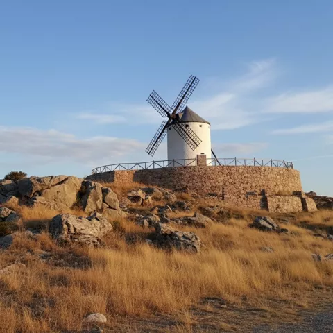 Molino de viento sobre base circular de piedra entre rocas y hierba seca, con cielo despejado.
