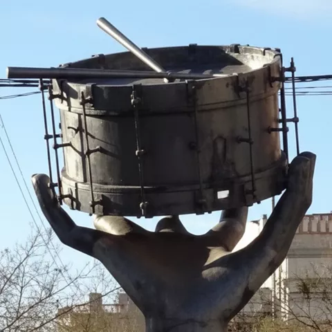 Primer plano de la escultura metálica con mano alzada y tambor, con edificios al fondo.