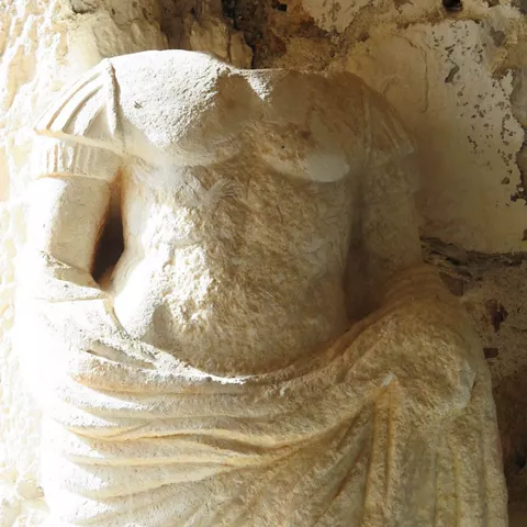 Escultura de piedra sin cabeza con manto tallado, situada en un espacio de muros antiguos.