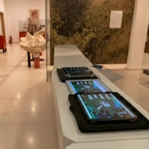 Mesa interactiva con pantallas táctiles en una sala de museo con vitrinas al fondo.
