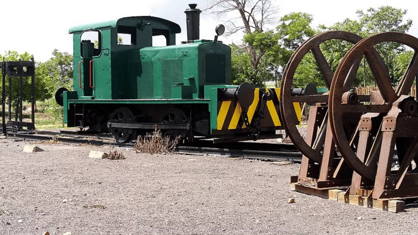 Locomotora minera verde sobre vías antiguas junto a grandes ruedas y maquinaria industrial