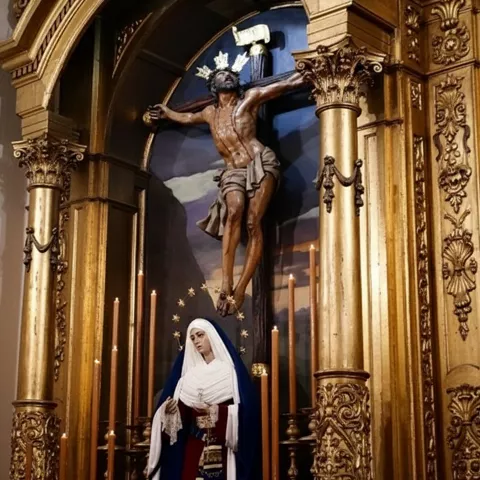 Altar ornamentado con columnas doradas y crucificado sobre fondo pintado, acompañado por imagen femenina y cirios encendidos.v