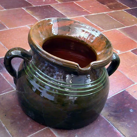 Recipiente de cerámica vidriada con dos asas y boca ancha sobre suelo de barro.