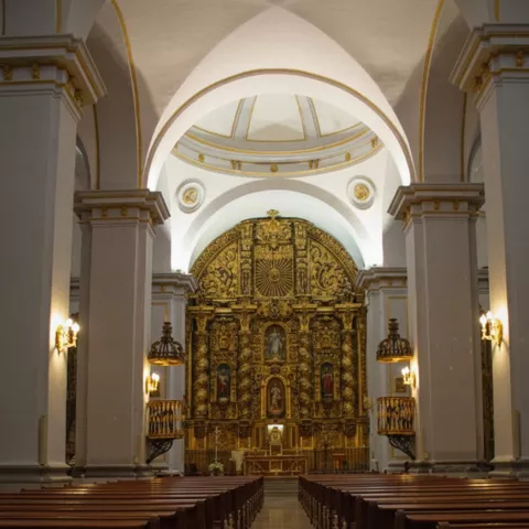 Nave central de iglesia con pasillo, bancos y retablo dorado al frente.