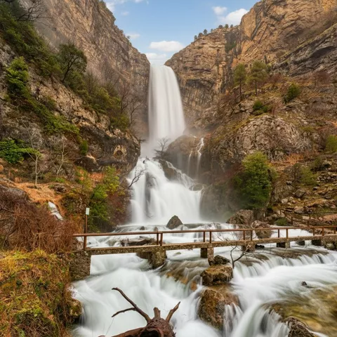 Cascada caudalosa entre paredes de roca, con pasarela de madera sobre el río.
