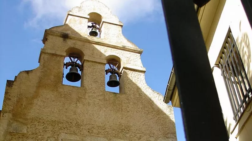 Fachada de edificio histórico con espadaña y campanas vista desde una calle estrecha.