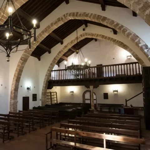 Nave interior con arcos de piedra, coro elevado y lámparas colgantes.