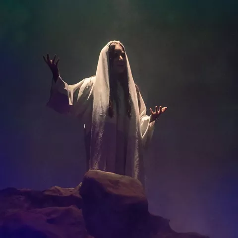 Figura vestida de blanco levanta los brazos bajo un haz de luz