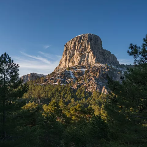 Peñón rocoso iluminado por el sol sobre un bosque de pinos con nieve dispersa.