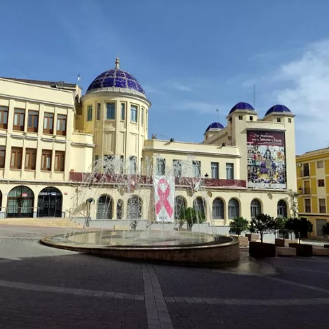 Plaza urbana con edificio histórico de fachada clara, cúpulas azules y fuente central en primer plano.