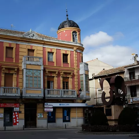 Vista de un edificio comercial con fachada de color rosa y amarillo y balcones de hierro forjado, situado en la esquina de una calle con un monumento de metal oxidado.