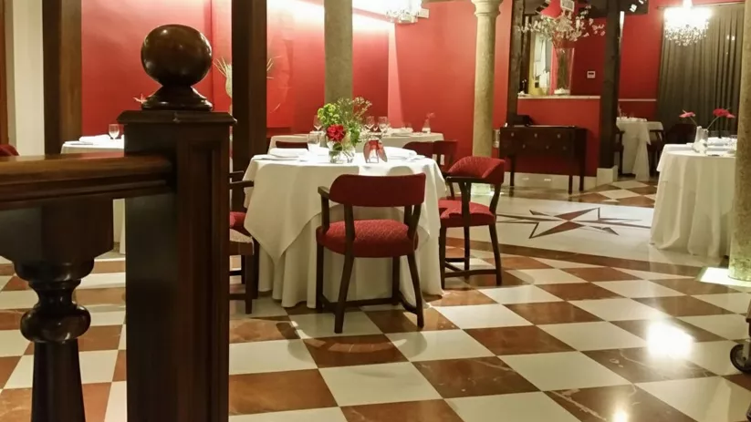 Restaurante con paredes rojas, suelo ajedrezado y mesas vestidas en blanco.
