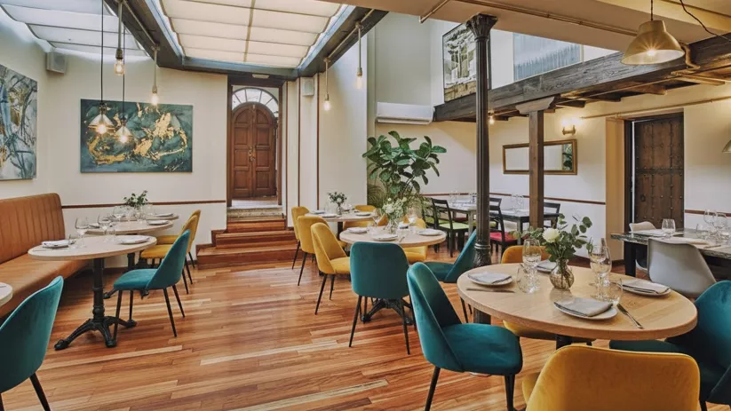 Sala luminosa con mesas redondas, sillas tapizadas en tonos mostaza y verde, y puerta de madera al fondo.