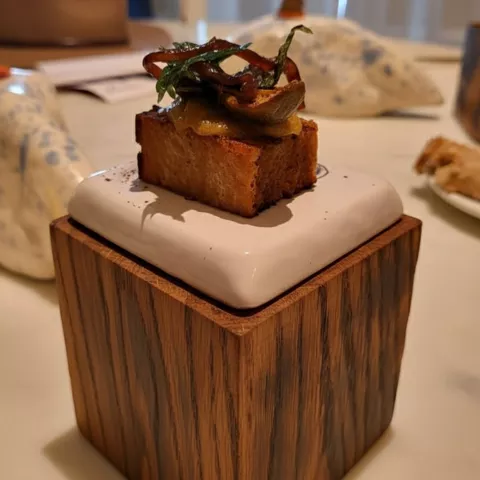 Bocado gourmet sobre base de madera con copa de vino desenfocada al fondo.