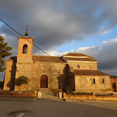 Fachada principal de una iglesia rural de piedra con una gran puerta arqueada de madera y una torre campanario con campana visible, capturada durante la luz dorada de la tarde con nubes en el cielo.