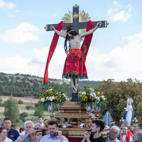 Paso con crucifijo adornado con flores, rodeado de personas durante una procesión al aire libre.