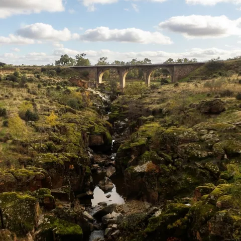 Paisaje de un valle profundo con formaciones rocosas cubiertas de musgo, un pequeño arroyo y un puente moderno al fondo.