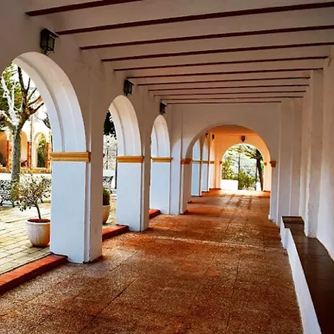 Galería cubierta con arcos blancos, suelo rojizo y luz natural al fondo.