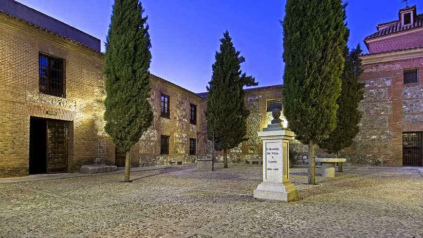 Patio empedrado con cipreses y busto sobre pedestal rodeado de edificios antiguos.