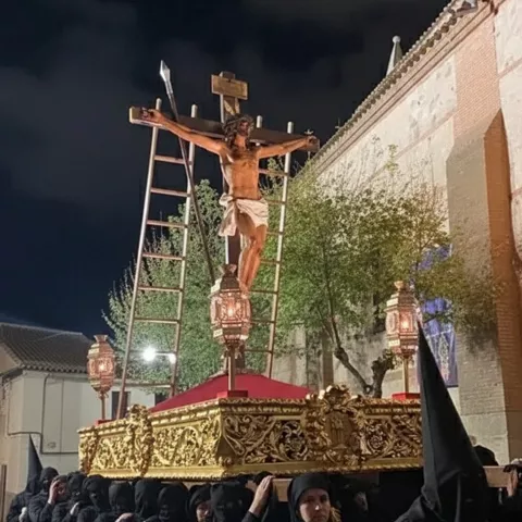 Paso nocturno con crucificado iluminado y fila de nazarenos con capirote negro.