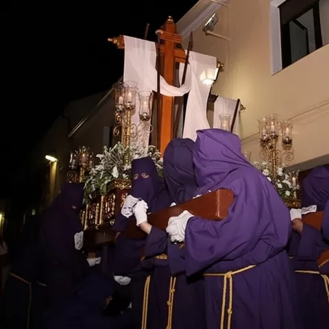 Cofrades con túnicas moradas portando un paso con cruz en procesión nocturna.