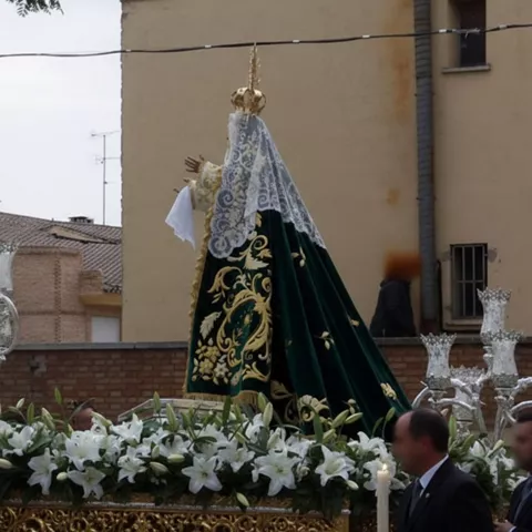 Imagen religiosa cubierta con manto verde bordado y velo blanco, sobre un paso con flores blancas y cirios.