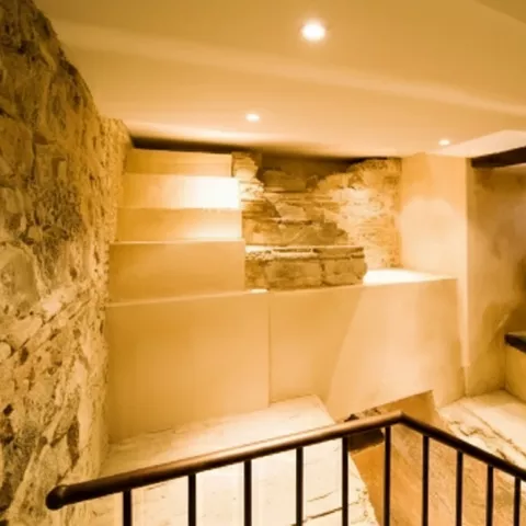 Escalera interior con muros de piedra y restos arqueológicos iluminados en sótano histórico.