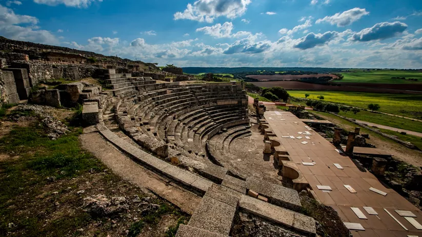 Gradas semicirculares de un teatro antiguo de piedra con paisaje rural.