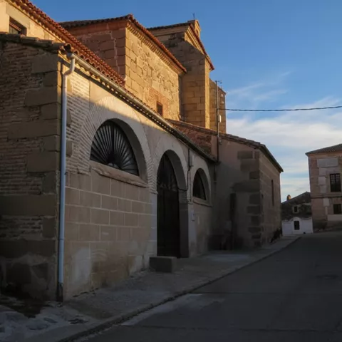 Perspectiva de una calle lateral donde se aprecia el lateral de una iglesia de piedra con grandes arcos cegados y un edificio tradicional al fondo bajo la luz del atardecer.