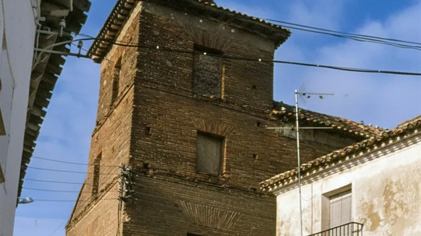 Una torre de ladrillo antiguo integrada en una calle estrecha entre casas de fachadas blancas y desgastadas.