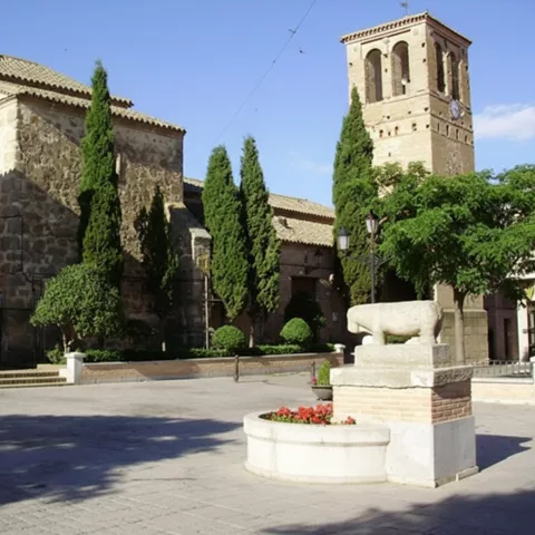 Fachada lateral de una iglesia de piedra y ladrillo con una torre campanario, árboles ornamentales y una escultura zoomorfa en primer plano.