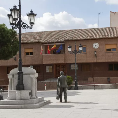 Una plaza pública con una fuente de piedra, estatuas de bronce y un edificio administrativo de ladrillo al fondo con banderas.