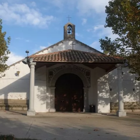 Una iglesia blanca con techo de tejas rojas y un pequeño campanario sobre la entrada principal, rodeada de frondosos árboles verdes.