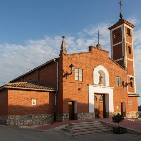 Perspectiva lateral de una iglesia de ladrillo bajo la luz del atardecer, destacando su zócalo de piedra y la torre campanario al fondo.