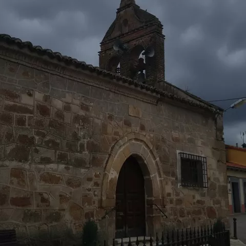 Fachada lateral de una pequeña iglesia de piedra con un arco de entrada apuntado y una espadaña de ladrillo con campanas, bajo un cielo oscuro y tormentoso.