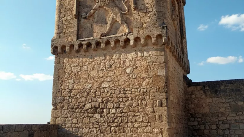 Torre de piedra de mampostería gruesa con un gran relieve tallado de un león rampante en su cara principal, bajo un cielo azul despejado.