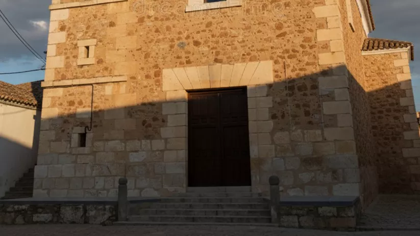 Una iglesia de piedra con un campanario cuadrado y un reloj bajo un cielo azul parcial.