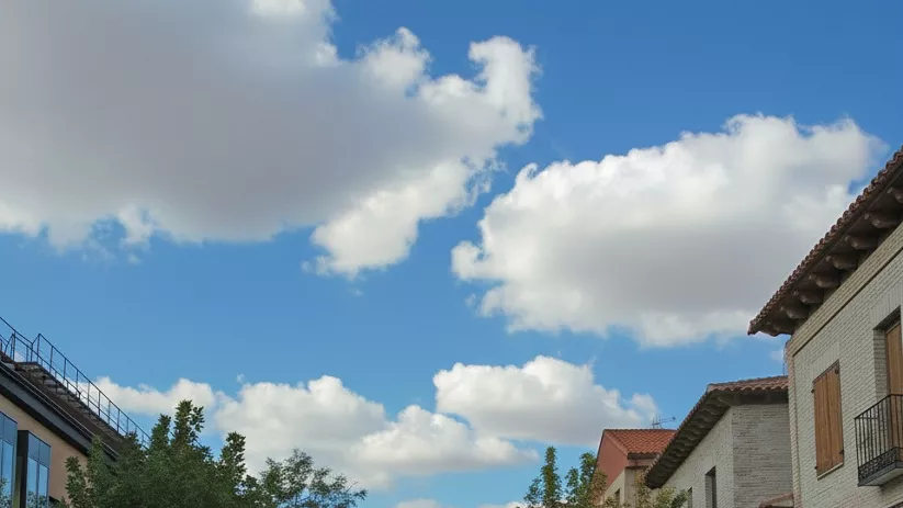 Una plaza de pueblo adoquinada con una fuente ornamental de varios niveles y un campanario de iglesia que se asoma detrás de los árboles.
