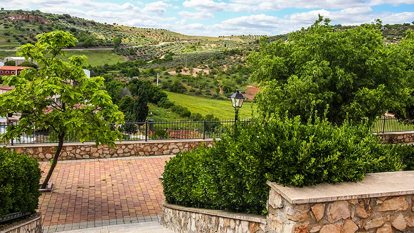 Vista panorámica de un paisaje rural con colinas verdes, árboles frondosos y un paseo empedrado con barandilla y farola, bajo un cielo azul con nubes.